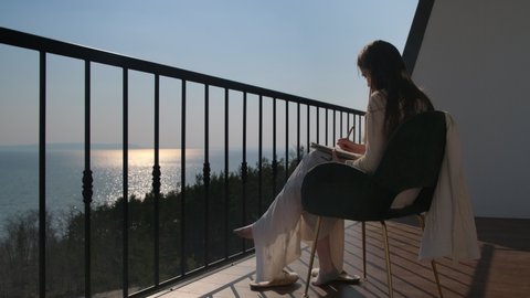 Inspired young girl illustrator posing alone in armchair near balcony railings drawing in her notepad. Amazing view of calm sea at dusk or dawn on background. Low angle shot, handheld