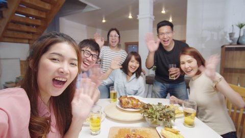 Group of young Asian people at home dinner party, looking at camera, greeting friends on remote video call. Point of view vlog or selfie camera. New normal lifestyle, social distancing concept