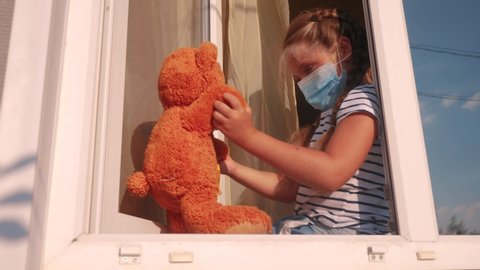 The bored kid in the medical mask in home quarantine coronavirus sitting covid 19 by the window. a child with a toy teddy bears in a protective mask looking out the window. coronavirus epidemic prevention concept.