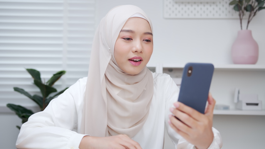 Beautiful Asian Muslim Woman in hijab making a video call smiling and talking with her friend and family. Businesswoman having video call discussing, working online meeting with team at home office Royalty-Free Stock Footage #1057351741