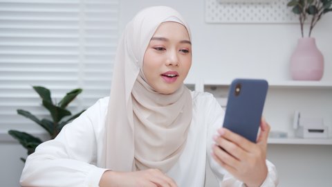Beautiful Asian Muslim Woman in hijab making a video call smiling and talking with her friend and family. Businesswoman having video call discussing, working online meeting with team at home office