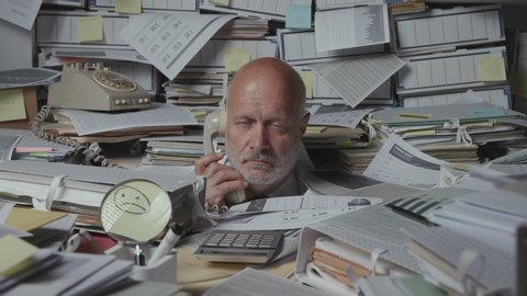 Sad frustrated business executive overwhelmed by work, he is overloaded with paperwork and answering phone calls in the office