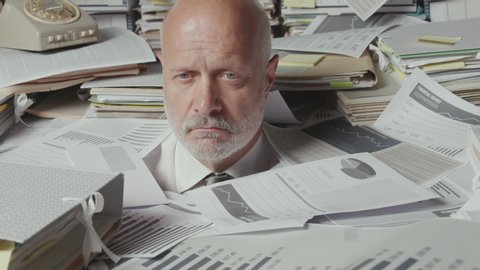 Sad stressed businessman working in his office, he is sitting under a lot of paperwork and he is overwhelmed by work
