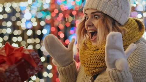 Portrait young woman receiving Christmas present from friend is surprised, smiles, picks up red box in her hands in warm mittens, rejoices at bokeh of Christmas lights in city square. Noel. Lifestyle