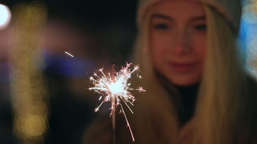 Christmas lights splashes burn in hands blonde girl smiling against of bokeh of Christmas lights in city square in knitted mittens hat winter. Sparklers. Noel. Christmas preparation. Emotions