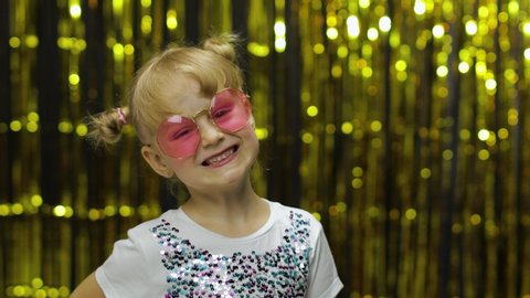 Child smiling, looking at camera. Little fun blonde kid teen teenager girl 4-5 years old in shiny white t-shirt, pink sunglasses posing isolated on background with foil fringe golden curtain in studio