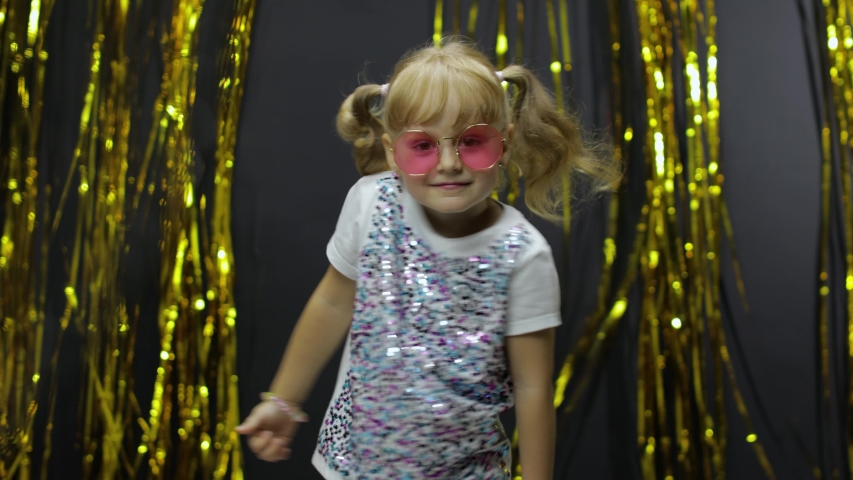 Stylish child dancing, make faces, shaking shoulders in silly dance. Little fun blonde kid teen girl 4-5 years old in pink sunglasses fooling around on background with foil fringe golden curtain Royalty-Free Stock Footage #1057357705