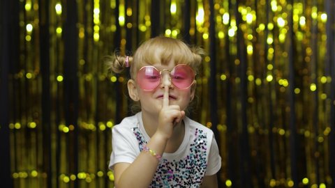 Child putting finger on lips making silence gesture, asking to keep secret. Shh. Be quite. Little fun blonde kid teen teenager girl 4-5 years old in sunglasses on background with foil fringe curtain