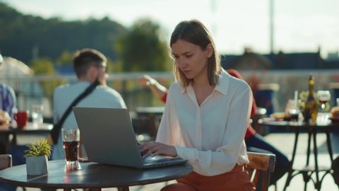Serious beautiful young woman sitting at table with laptop in cafe working online typing internet online communication technology business outdoors slow motion