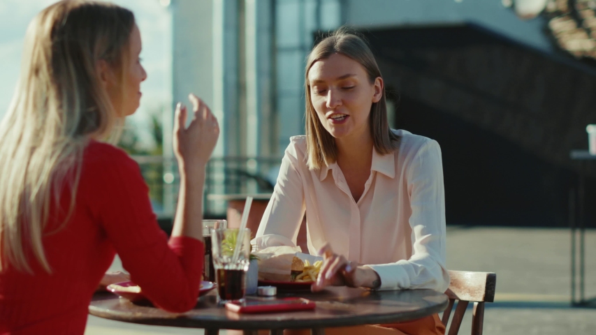 Two cheerful women resting talking drinking cocktail eating burger in summer cafe street sisters cafe casual city friendship models outdoors slow motion Royalty-Free Stock Footage #1057358929