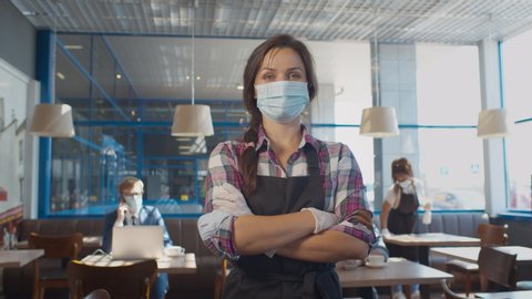 Portrait of waitress wearing face mask and gloves while serving in coffee shop. Pretty restaurant worker in apron and protective mask and gloves posing at camera with crossed hands