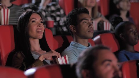Close-up of attractive smiling young couple chatting while watching movie at cinema theater, beautiful woman eating popcorn, man drinking coda. Multinational audience enjoying interesting movie