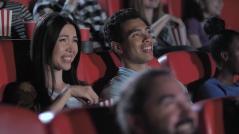 Closeup of nice asian couple enjoying comedy in movie theater, cute brunette female eating popcorn and bursting from laughter together with boyfriend. Laughing diverse viewers watching movie in cinema