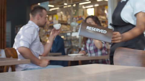 Close up of waiter putting reserved plate on table in restaurant with happy couple drinking coffee on date on blurred background. Restaurant manager placing reserved table sign
