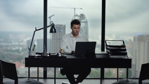 Focused businessman working on laptop computer in office with large windows. Male worker using laptop computer at remote workplace. Serious business man looking at laptop screen in home office