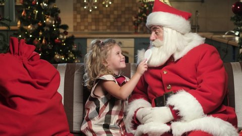 little Girl on Christmas night is sitting on the sofa and combing Santa Claus's big fluffy white beard. Santa with kid at home during the Christmas holidays. Taking care of Santa in the new year.