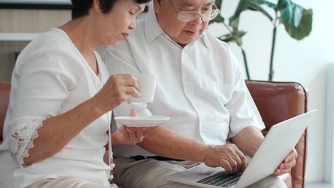 Happy Asian senior elderly couple using laptop talking together doing online shopping, senior mature retired family reading discussing internet computer news, choosing travel offer on website at home.
