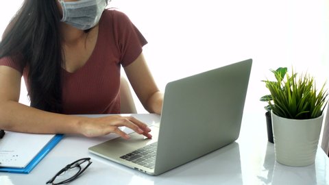 Asian business woman wear face mask protection work from home she is video conferences by modern laptop computer with the team and use hand with pencil writing on paper book note inspire idea