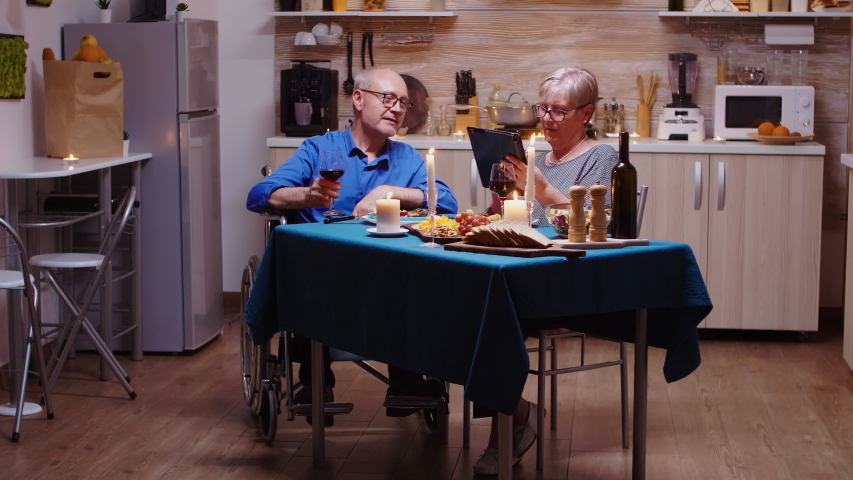 Elderly retired woman showing tablet to paralyzed man, scrolling and showing photos. Imobilized handicapped senior husband scolling on phone enjoying the festive meal.