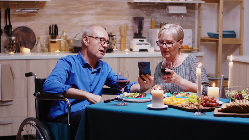 Man in whellchair using smartphone sitting at the table in the kitchen while romantic dinner. Imobilized paralyzed handicapped elderly husband scolling on phone enjoying the festive male.