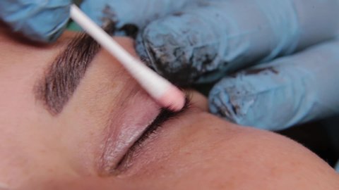 beautician is using small cotton swab for remove dye from eyelids of female client in a beauty salon.