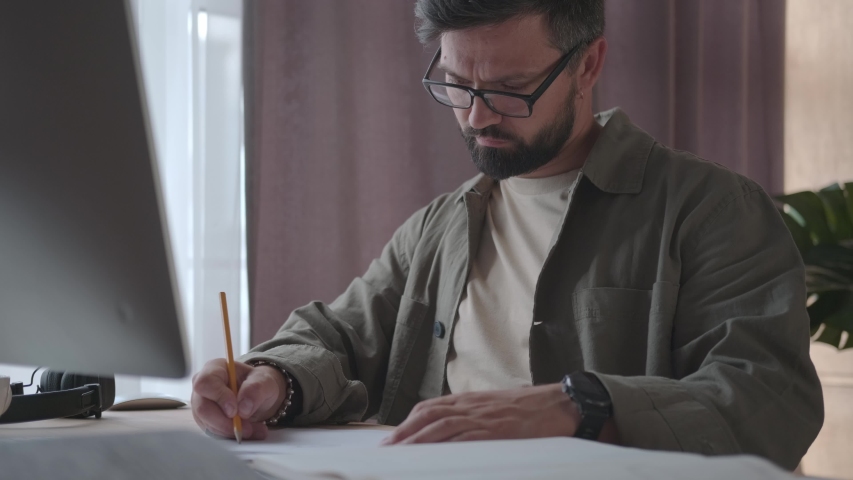 A serious man with glasses with a beard is developing a project. He works with papers, draws and paints sitting at the desk in a neutral office interior. Medium shot. Remote work. | Shutterstock HD Video #1057384048