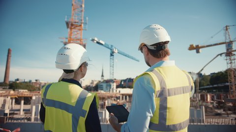 Male Civil Engineer and Young Female Building Architect Use a Tablet Computer on a City Construction Site. They Talk About the Future of Real Estate Development and Planning. Wearing Safety Hard Hats.