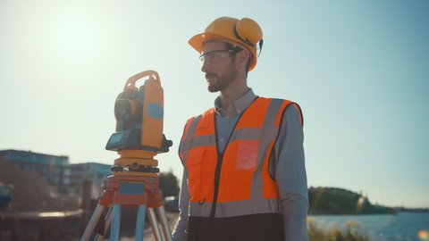 Construction Worker Using Theodolite Surveying Optical Instrument for Measuring Angles in Horizontal and Vertical Planes on Construction Site. Worker in Hard Hat Making Projections for the Building.