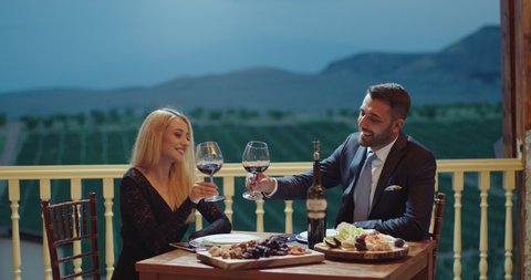Young couple dining exclusively in a romantic atmosphere in restaurant . Pair enjoying romantic dinner in terrace near the vineyard . Couple drinking wine and making toast . Slow Motion .