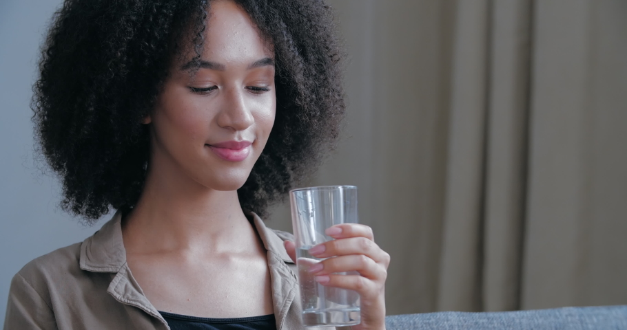 Close up of young African American woman quenches her thirst, holds glass cool warm water, drinks, closes his eyes in pleasure, smiling looks at camera. Attractive curly haired girl freshening up Royalty-Free Stock Footage #1057393114
