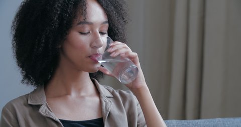 Close up of young African American woman quenches her thirst, holds glass cool warm water, drinks, closes his eyes in pleasure, smiling looks at camera. Attractive curly haired girl freshening up