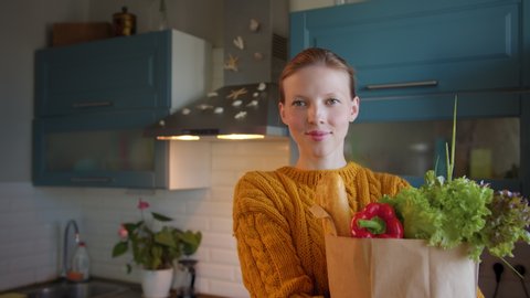 Young beautiful woman with groceries bag in hands at kitchen looks camera smiling and happy. Home delivery products. Buying fresh food, conscious consumption