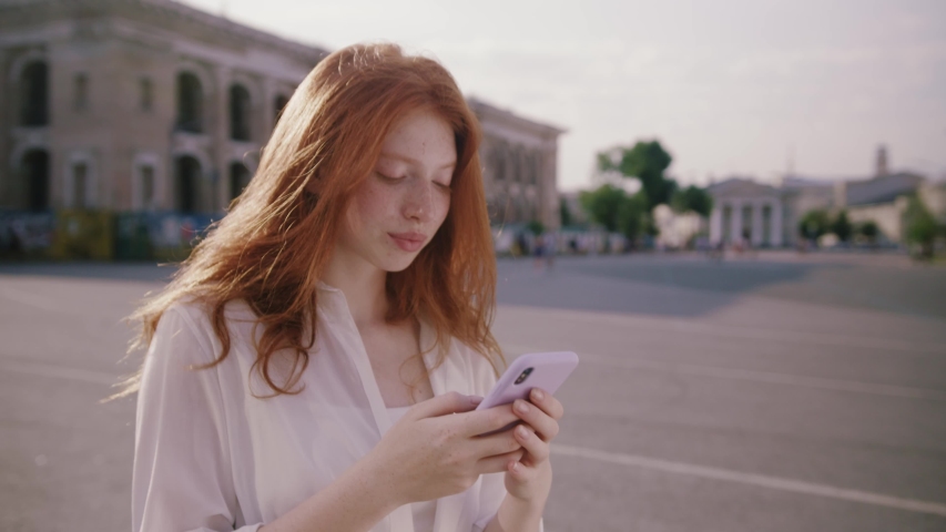 Smiling face young woman with red hair use phone walking in the city streets looking around feel happy portrait technology cellphone mobile pretty cityscape slow motion