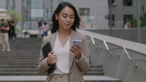Smiling asian woman wearing formal suit using mobile phone typing text messages walking holding laptop outside business cellphone city smartphone slow motion
