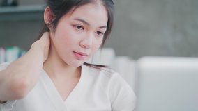 Asian woman tired while sits at a work desk at home office. Young Asian businesswoman self-massaging with a painful face. Concept of new normal lifestyle, remote working, online learning at home.