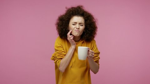Dental problems. Girl afro hairstyle in hoodie drinking hot or ice water and feeling sudden terrible toothache, pain from sensitive tooth and cavities. indoor studio shot isolated on pink background