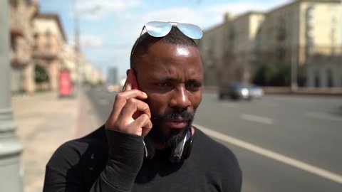 Portrait of a nervous African-American man using a mobile phone on a city street against a highway background. He has wireless headphones around his neck.