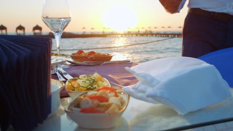 The waiter drops the fish dish to the dining table set up on the pier. sunset. seaside. slow motion.