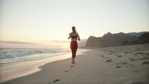 Woman athlete running away into sunset at beach of California, running workout outside, burgundy sport suit, view from back