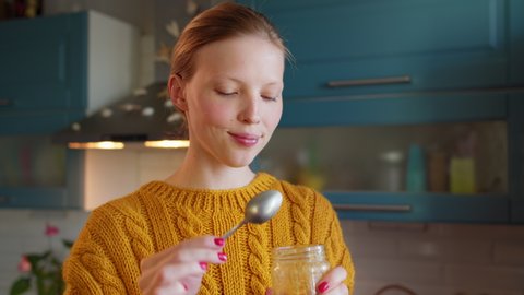 Close up young woman in kitchen eating honey from jar with spoon and enjoying meal. Attractive woman eating healthy food spoon