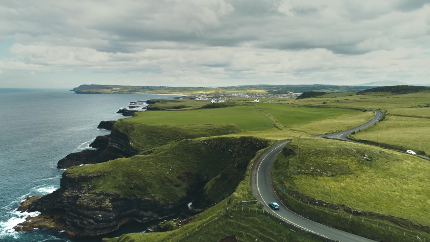 Green fields, meadows aerial: road cars driving on background. Ireland's hills, farmlands, clouds on horizon. Picturesque landscape view of Antrim County, United Kingdom. Footage shot in 4K, UHD Royalty-Free Stock Footage #1057416877