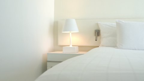 In a white hotel room, there is a white lamp on a white bedstand illuminating a headboard and the crisp white linen of a perfectly made-up bed.