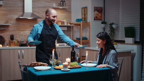 Man preparing festive dinner for his beautiful wife. Young couple talking, sitting at the table in kitchen, enjoying the meal, celebrating their anniversary at home with healty food. Romantic happy