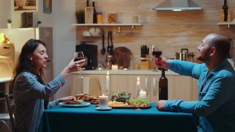 Loving young couple raising red wine glass and toasting while enjoying romantic dinner at home in the kitchen. Happy in love people eating meal, celebrating anniversary in dining room, romantic toast Arkivvideo