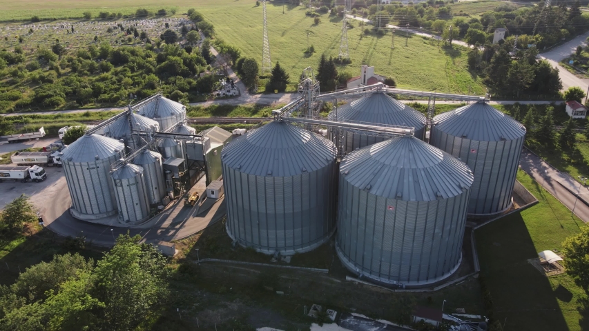 Farm Grain Storage Bins wheat field at sunset from top aerial view. Steel Granary storage tanks steel warehouse granary for storage of loose grain blue sky volumetric clouds. Elevator. Parked trucks.