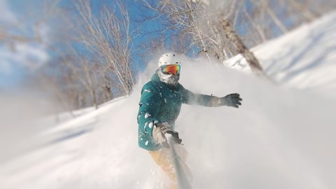 Man riding on snowboard with selfie stick in his hand between trees on slow motion. Guy doing snow splash rising on magnificent sunny day. Concept of extreme, sport, winter, freeride, snowboarding