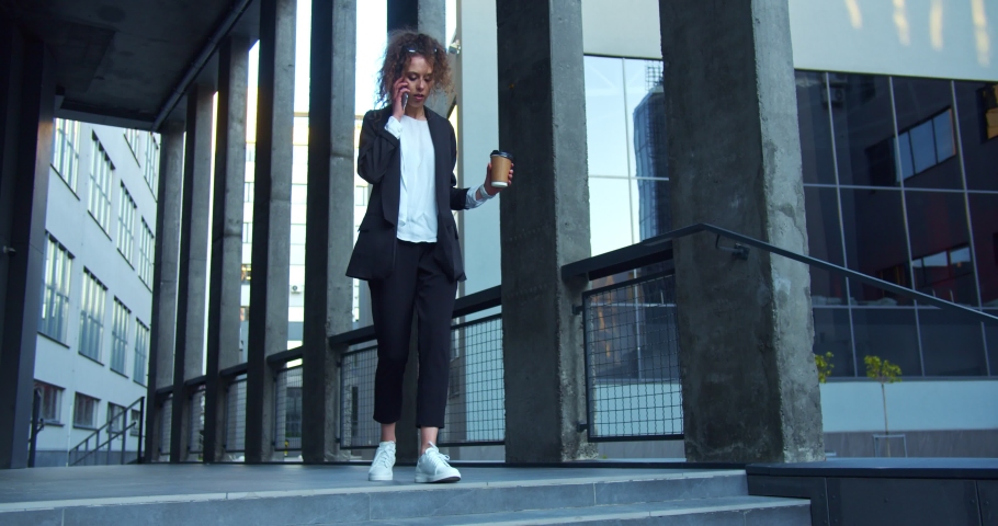 Hurrying Caucasian businesswoman in a formal suit with sunglasses. Busy female worker rushing to the meeting and spilling the coffee cup while talking on the phone. Modern scyscrapers background. Royalty-Free Stock Footage #1057431910