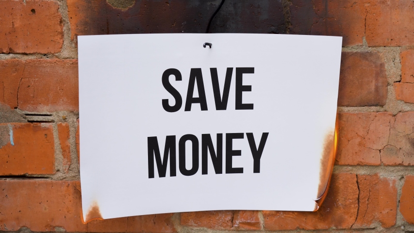 The inscription on a white A4 size sheet against the background of an orange brick wall. The paper with the black word Save Money burns, smoke and turns into ashes. Business concept.