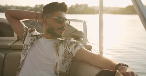 Handsome stylish young hindu guy in sunglasses in white t-shirt and shirt sits on a yacht or boat.