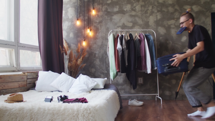 Be late guy with tickets in his teeth puts a suitcase on the bed, shoves things with hangers and a camera into it, against the background of the apartment. Rushing guy, quickly collects things in a